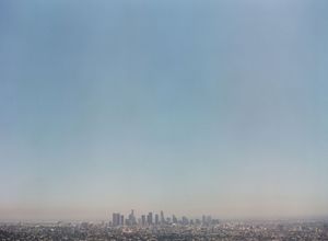 Hazy aerial view of Los Angeles downtown skyline stretching across the horizon under a pale blue smoggy sky.