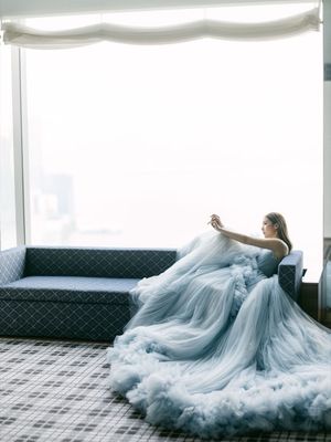 Bride in a dramatic flowing blue tulle gown seated by a large window in a luxury hotel.
