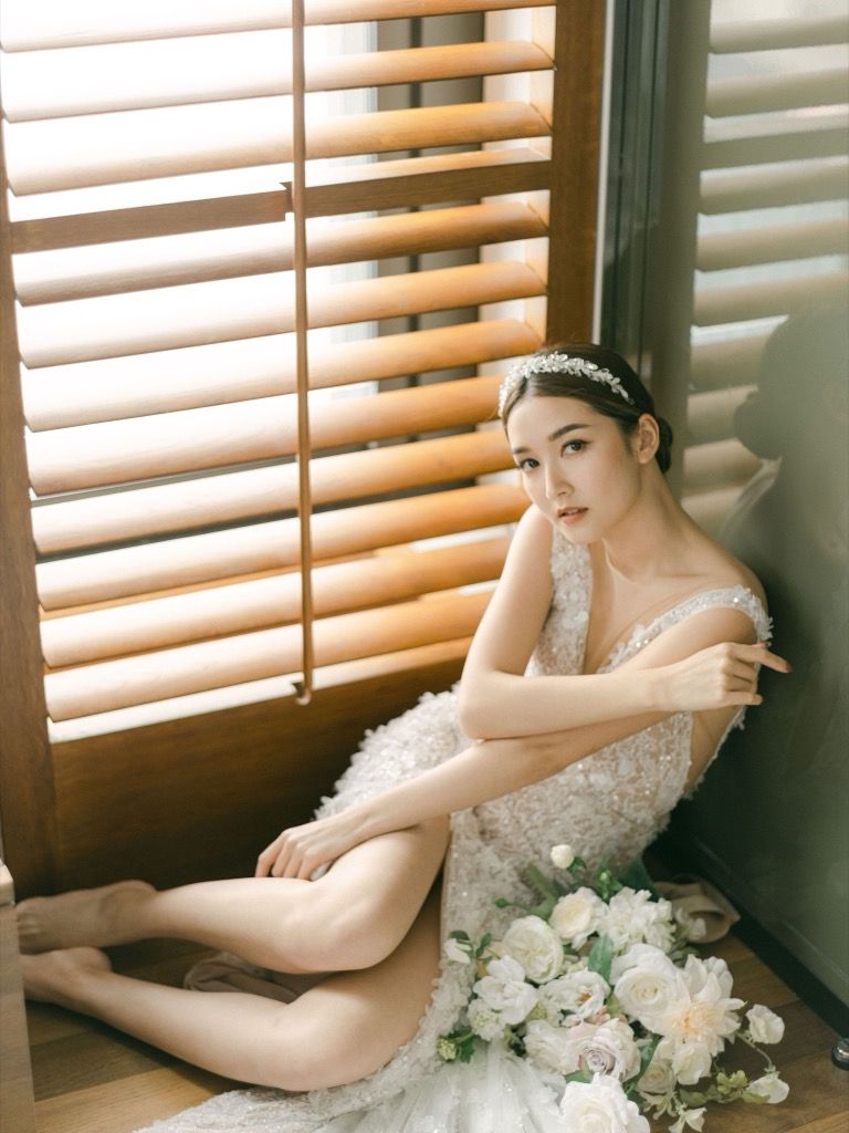 Bride in a lace gown sitting by wooden shuttered window with white roses bouquet.
