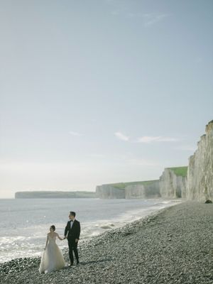 Bride and groom holding hands on a rocky beach with dramatic white chalk cliffs in the background.