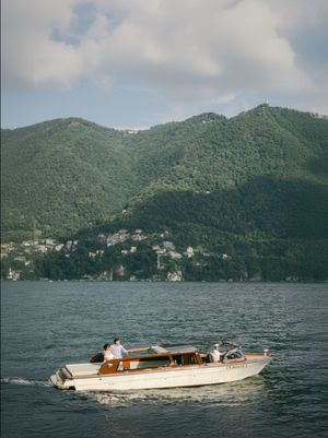 Couple riding a classic wooden speedboat on Lake Como with lush green mountains behind.