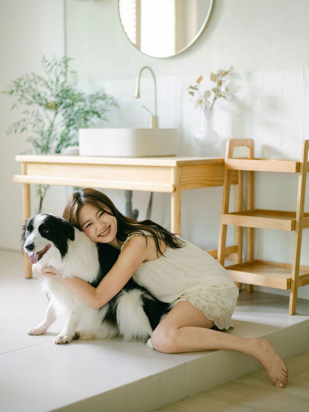 Smiling woman hugs black and white border collie dog on floor of bright modern bathroom.