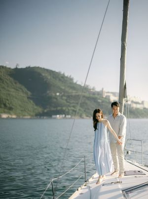 Couple holding hands on sailboat bow with green hillside and calm blue water backdrop.