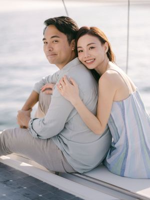 Couple sitting on sailboat deck, woman smiling and embracing man from behind over open sea.