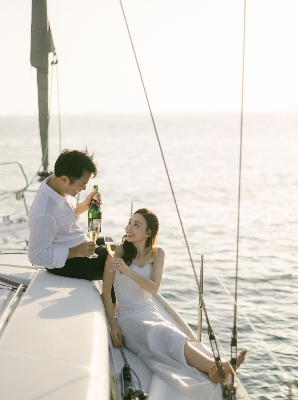 Couple toasting with bottles on sailboat deck at golden sunset over calm ocean waters.
