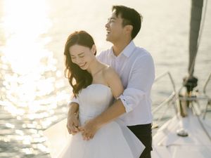 Bride in white gown embraced by groom on sailboat with sparkling sunlit ocean behind them.