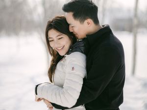 Smiling Asian couple embracing warmly in a snowy winter landscape with bare white trees.