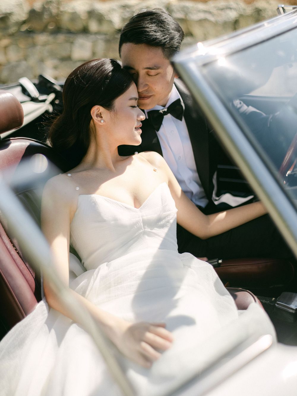 Bride in strapless gown and groom in tuxedo sharing intimate moment inside vintage convertible car.