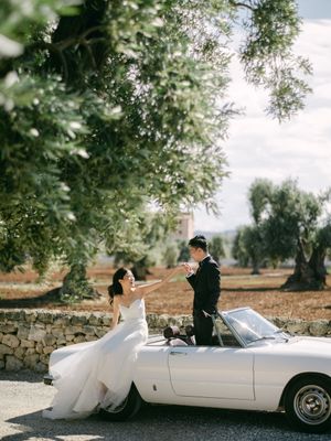 Bride and groom posing playfully with white vintage convertible under ancient olive trees in Italy.