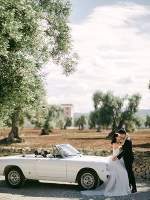 Bride and groom kissing beside white vintage convertible car surrounded by ancient Italian olive trees.