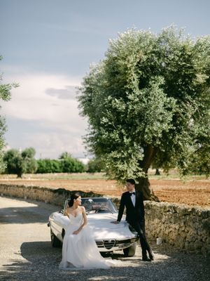 Bride and groom pose beside a vintage white convertible on a rustic Italian countryside road lined with olive trees.