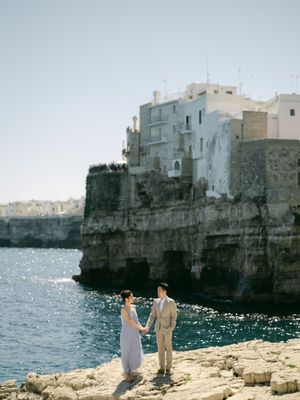 Couple holding hands on rocky coastline with stunning whitewashed cliffside buildings behind them.