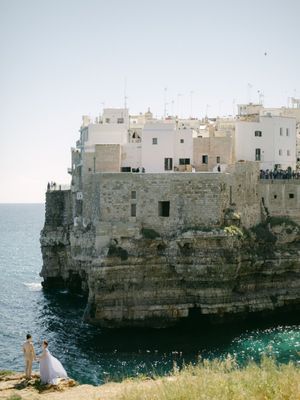 Couple in wedding attire standing before dramatic cliffside white buildings of Polignano a Mare.