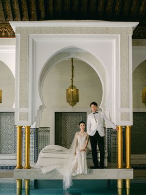 Elegant bride and groom pose beneath ornate Moroccan arches with gold columns beside an indoor pool.