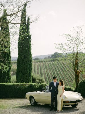 Bride and groom stand beside a vintage cream convertible with Tuscan vineyards and cypress trees behind them.