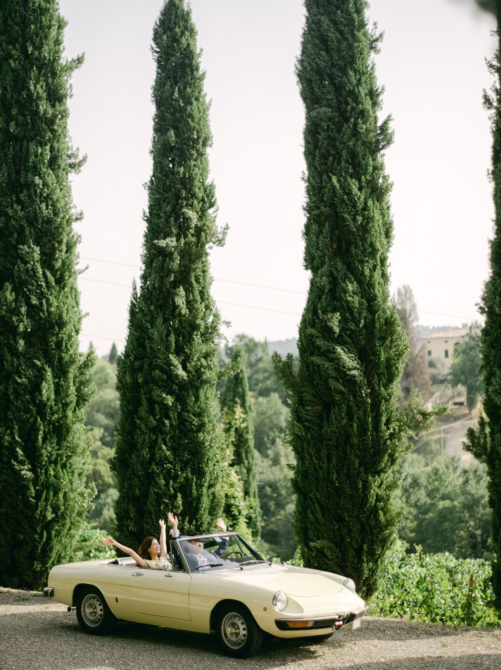 Couple rides in a vintage cream convertible convertible along a scenic road lined with tall Italian cypress trees.