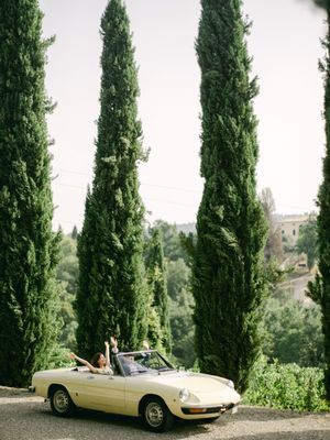 Couple rides in a vintage cream convertible convertible along a scenic road lined with tall Italian cypress trees.