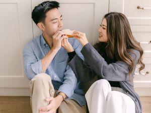 A happy couple sits on the floor sharing food in a cozy kitchen setting.