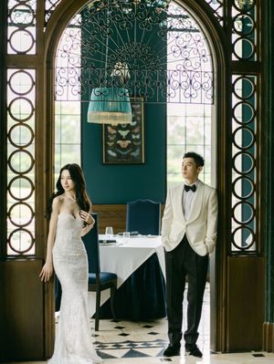 A bride in a lace gown and groom in a white tuxedo pose in an ornate restaurant doorway.