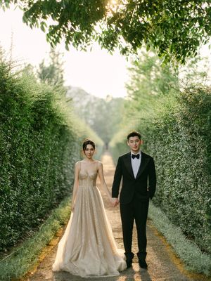 A couple in wedding attire holds hands on a tree-lined garden path at golden hour.