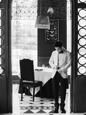 A groom in a white tuxedo adjusts his cufflinks in an elegant restaurant in black and white.