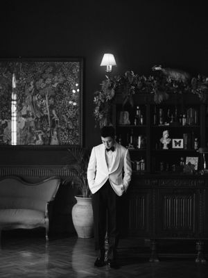 A groom in a white tuxedo stands pensively in a dimly lit elegant room in black and white.