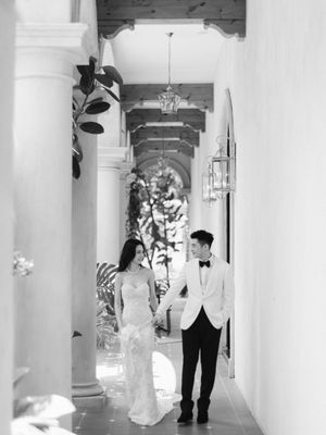A bride and groom walk hand in hand through a sunlit columned corridor in black and white.