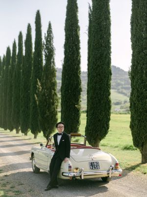 Groom in tuxedo leans against vintage convertible on cypress-lined driveway in Tuscany, Italy.