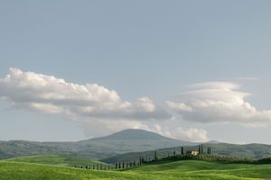 Rolling green Tuscan hills with cypress trees, farmhouse, and dramatic clouds over mountain landscape.