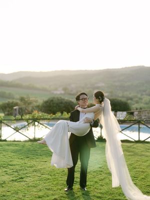 Groom carries bride in wedding dress at golden sunset with Tuscan hills and pool backdrop.