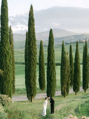 Bride and groom share a kiss among tall Italian cypress trees with rolling green Tuscan hills.