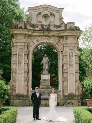 Bride and groom pose before ornate baroque stone arch with statue in elegant Italian garden wedding.