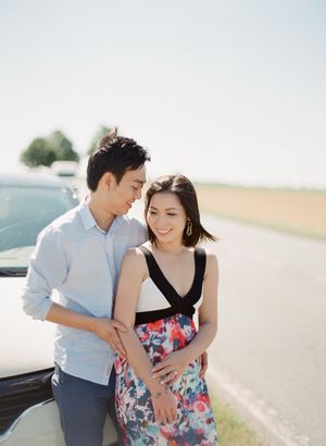 Asian couple leans against a vintage car on an open road, woman in colorful floral dress smiling warmly.