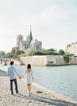 Couple walks hand in hand along the Seine River cobblestone bank with Notre-Dame Cathedral in the background.