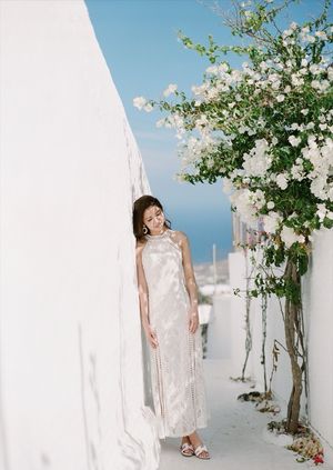 Bride in elegant white halter gown posing beside blooming white flowers on a whitewashed Santorini alley.