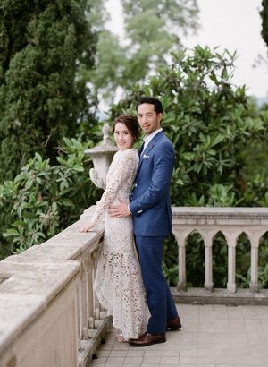 Couple posing on a stone balcony surrounded by lush greenery in a romantic European garden setting.