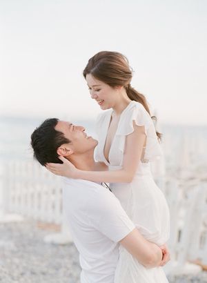 Man joyfully lifting smiling woman in white dress near a bright seaside fence at golden hour.