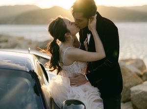 Bride and groom sharing a romantic sunset kiss leaning against a car by the waterfront.