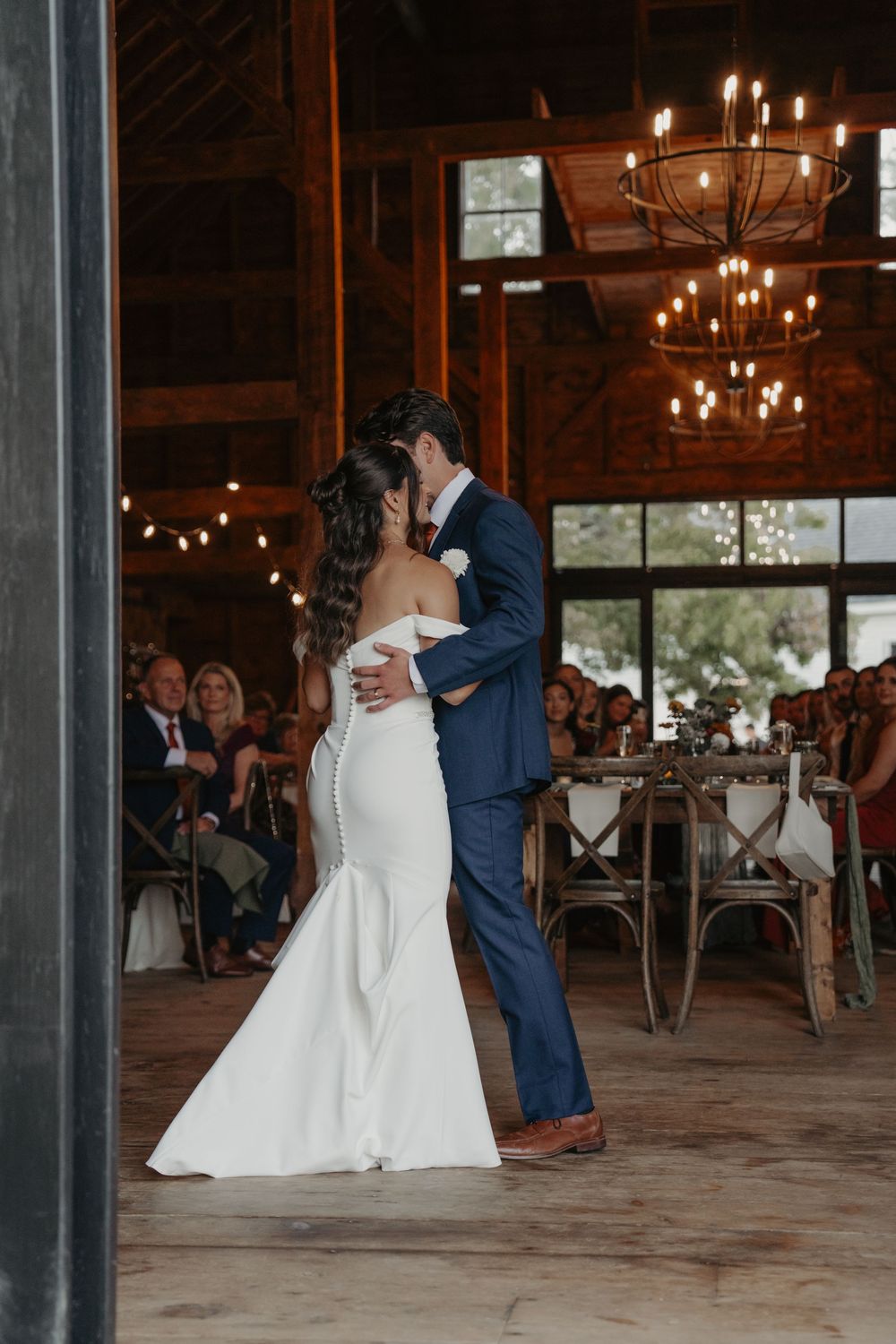 Newlyweds share their first dance in a rustic barn venue with chandeliers and string lights overhead.