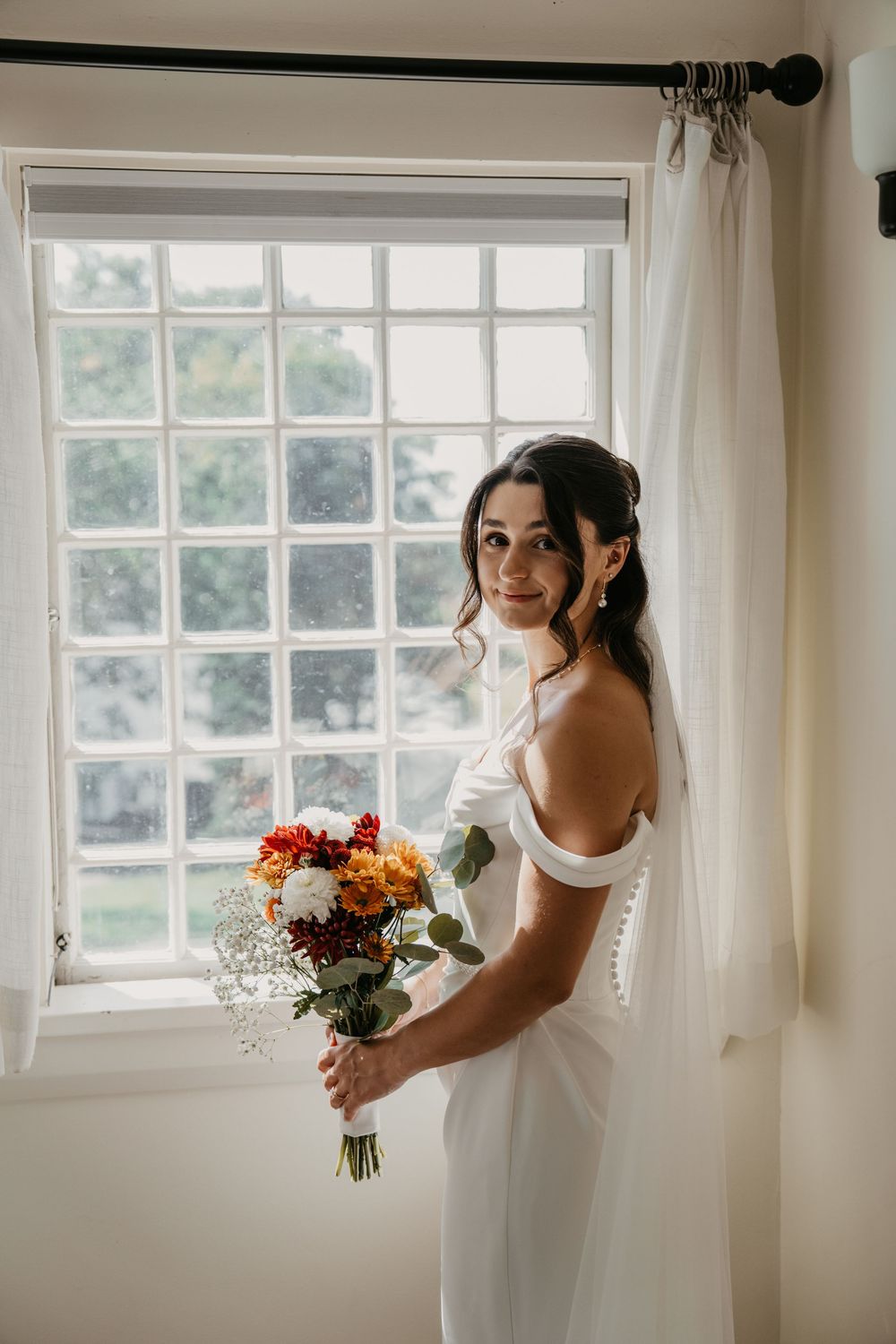 A bride in a white dress holds a vibrant autumn bouquet near sunlit windows in a bright room.