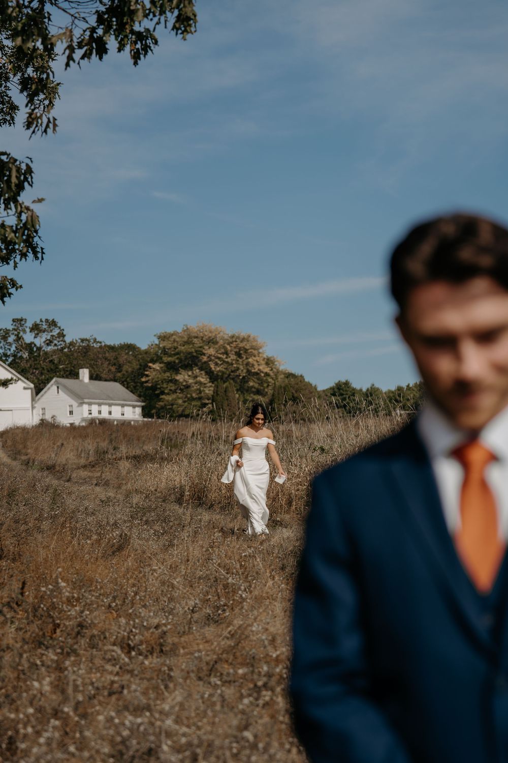 A wedding couple poses in a rustic field with a white farmhouse visible in the background on a sunny day.