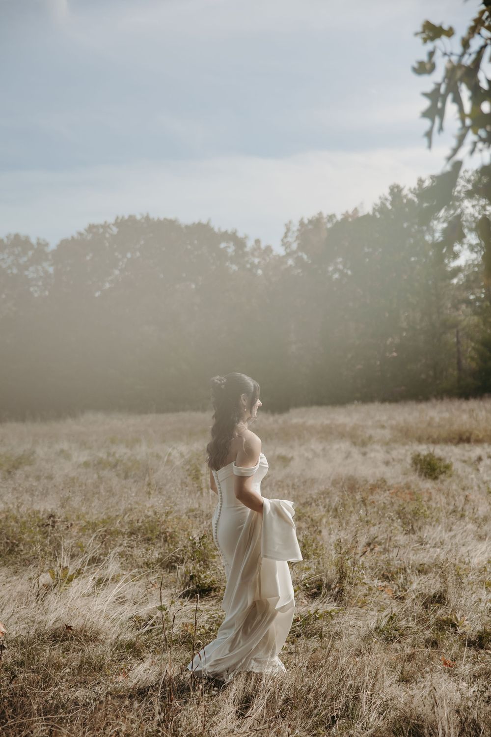 A bride in a flowing white dress walks through a sunlit meadow during golden hour.