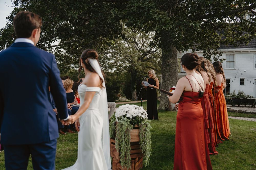Wedding party stands in a garden setting wearing navy and rust-colored attire during an outdoor ceremony.
