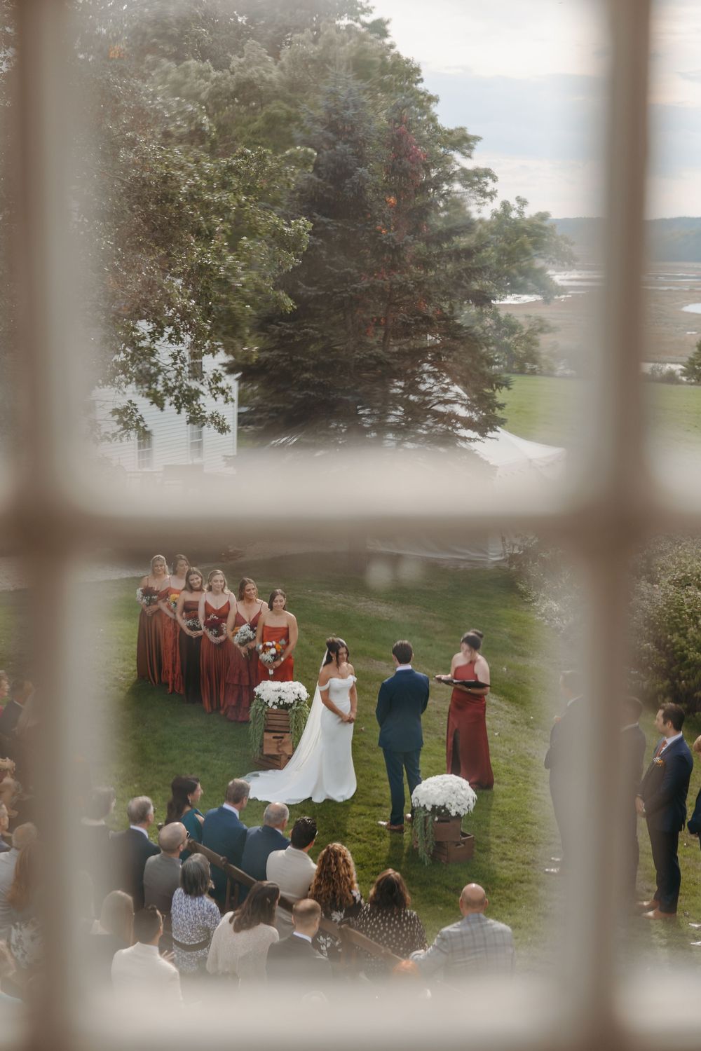 An outdoor wedding ceremony viewed through window panes with guests gathered on a grassy hillside.