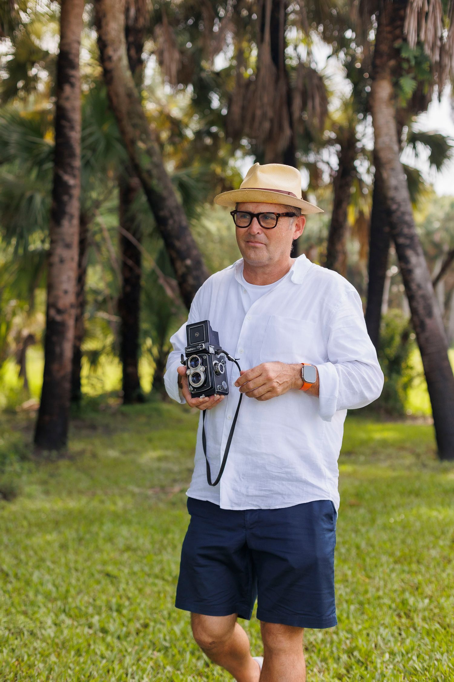 Man in white shirt and hat holds vintage camera in wooded outdoor setting.