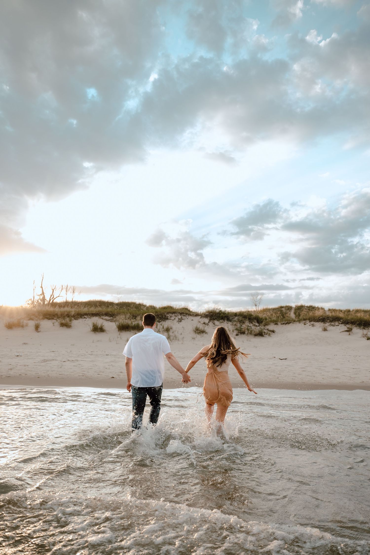 A couple runs into the surf at sunset holding hands on a sandy beach with dramatic cloudy sky above.