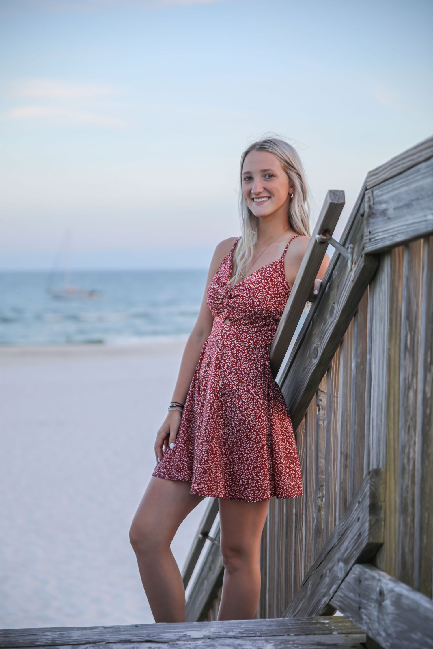 A person in a red floral dress poses against a wooden beach boardwalk railing at dusk.