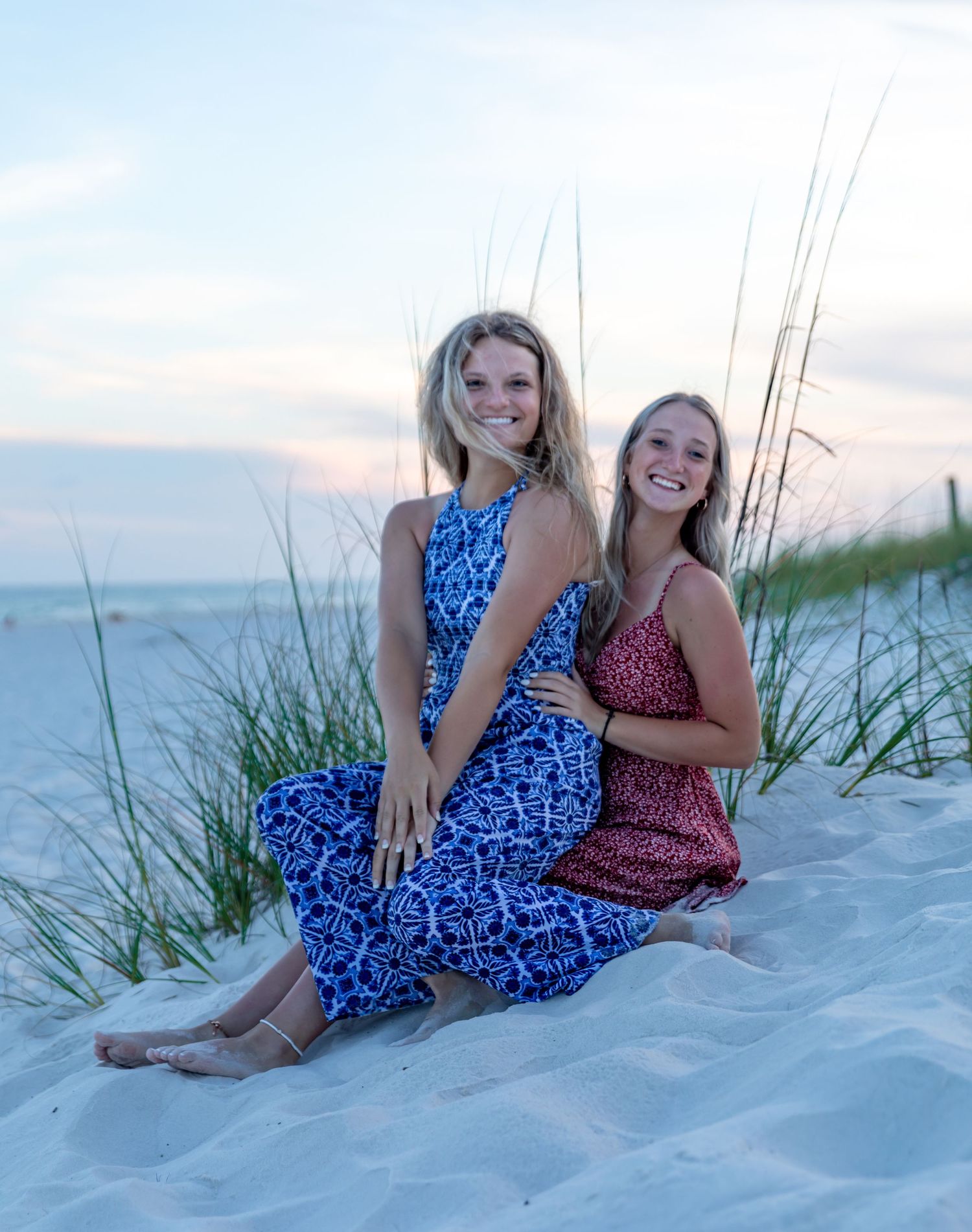 Two people in patterned dresses sit together on white sand dunes at sunset.