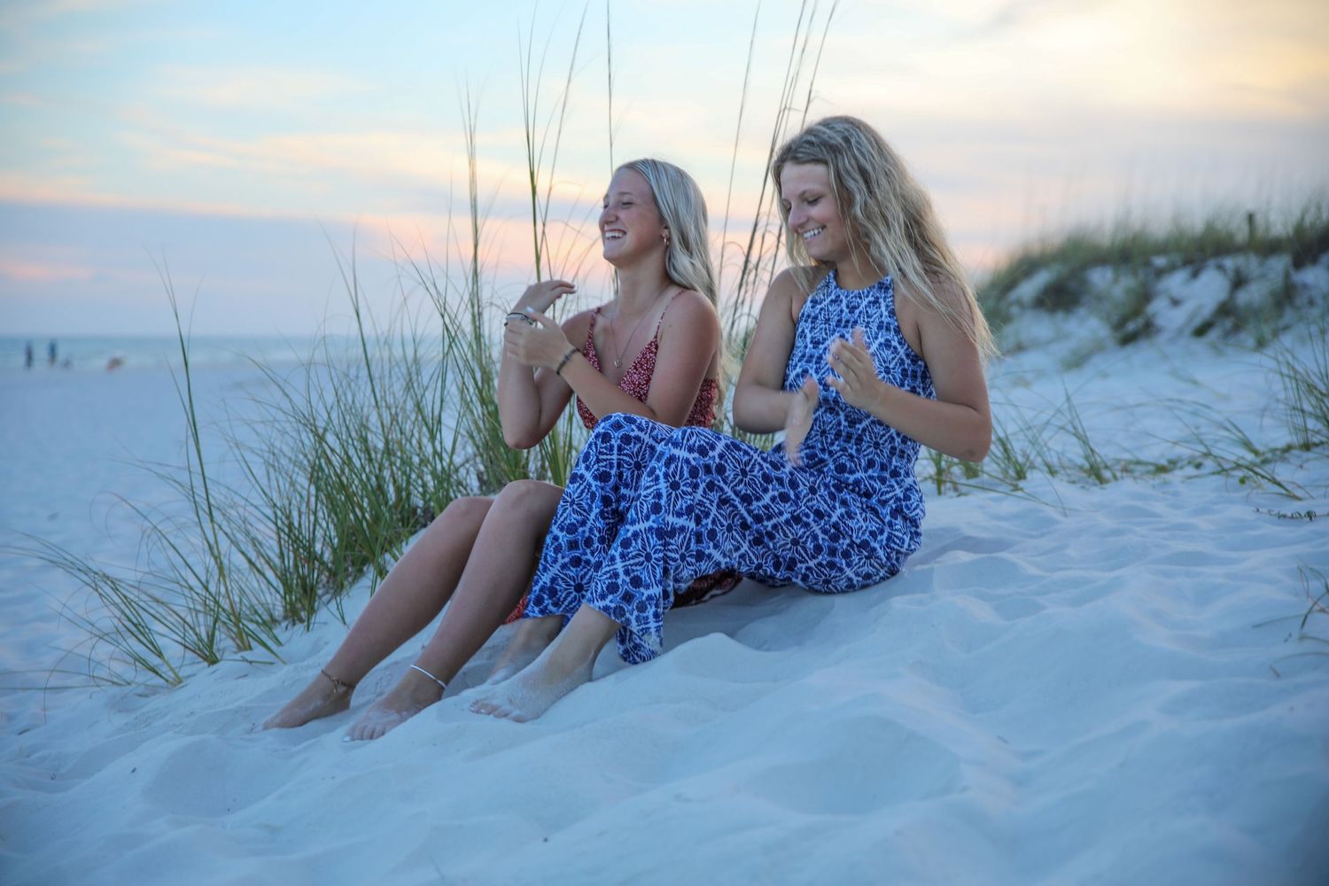 Two friends in summer dresses relax on sandy beach dunes during sunset.