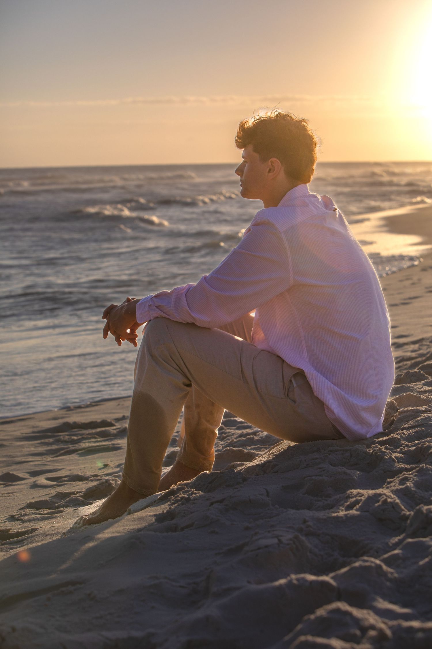 A silhouette sits on a sandy beach watching the sunset over ocean waves while wearing a white hoodie.